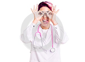 Young beautiful woman with pink hair wearing doctor uniform doing ok gesture like binoculars sticking tongue out, eyes looking