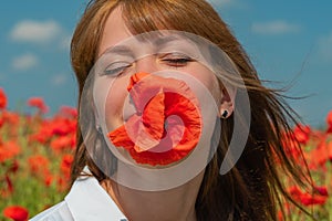 Young beautiful woman with a poppy flower in her mouth on a poppy field in summer.