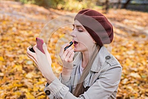 Young beautiful woman put on lipstick and looking in mirror on autumn Park background