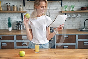 Young beautiful woman is reading her morning press and smiling while drimking orange juice in the kitchen. wearing white shirt