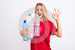 Young beautiful woman recycling plastic bottles standing over isolated white background doing ok sign with fingers, excellent