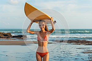 Young beautiful woman in red bikini and sun glasses holds in hands a surf on the ocean beach at sunset.