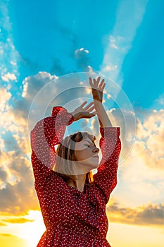 Young beautiful woman in a red dress against a blue sky with bright beams of the sun. The girl raised her hands elegantly