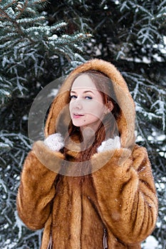 Young beautiful woman with red hair and blue eyes in faux fur coat is walking in winter snowy park