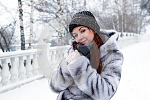 Young beautiful woman with red hair and blue eyes in gray faux fur coat with hood is walking in winter snowy park