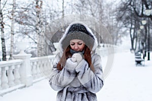 Young beautiful woman with red hair in gray faux fur coat with hood on background of winter snowy park