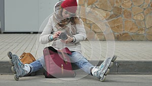 Young beautiful woman in red hat wearing sporty warm clothes and rollers, sitting on the asphalt road and taking