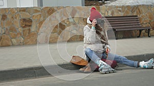 Young beautiful woman in red hat wearing sporty warm clothes and rollers, sitting on the asphalt road and taking