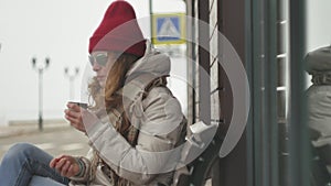 Young beautiful woman in a red hat wearing sporty warm clothes and rollers, sitting on a wooden bench drinking tea from