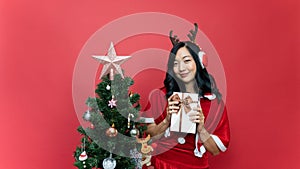 Young beautiful woman with red santy costume holding gift box. The scene has a Christmas tree and a red background