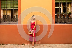 young beautiful woman in a red silk dress holds a pink handbag in her hands. The woman is leaning against a red and yellow wall in