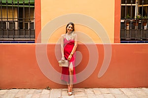 young beautiful woman in a red silk dress holds a pink handbag in her hands. The woman is leaning against a red and yellow wall in