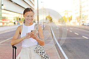 young beautiful woman with red suitcase is using smartphone while waiting for a taxi cab in the city. stylish smiling