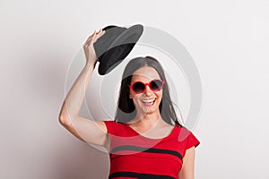 A young beautiful woman with red sunglasses lifting black hat in a studio.