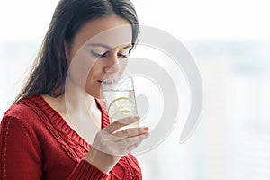 Young beautiful woman in red sweater with glass of water with lime, woman stands near window in skyscraper on cloudy day. Healthy