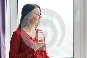 Young beautiful woman in red sweater with glass of water with lime, woman stands near window in skyscraper on cloudy day. Healthy