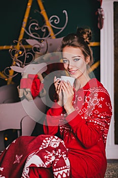 Young beautiful woman in a red warm pajamas with scandinavian ornaments sitting near decorative fireplace and drinking hot tea