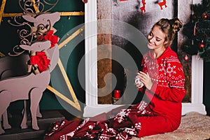 Young beautiful woman in a red warm pajamas with scandinavian ornaments sitting near decorative fireplace and drinking hot tea