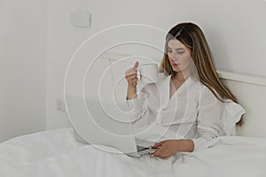 Young beautiful woman relaxing and drinking cup of hot coffee or tea using laptop computer on the bed in the bedroom.