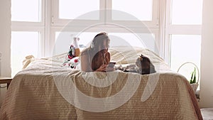 Young beautiful woman relaxing with her lovely Maine Coon cats laying in bed uses mobile phone and listen to music in