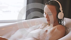 Young beautiful woman relaxing while listening to music in wireless headphones in bubble bath.