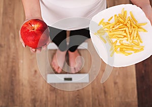 A young beautiful woman is on the scales and make a choice between an apple and potato chips. The concept of healthy eating. Healt