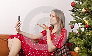 Young beautiful woman sending kiss while chatting by video call on mobile phone, sitting on sofa in pygamas near decorated xmas