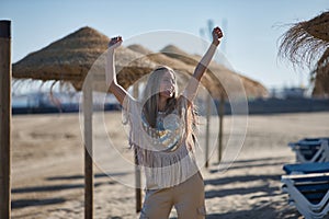 Young and beautiful woman with sequined shirt and bangs, with arms up, happy and smiling, between umbrellas and hammocks on the