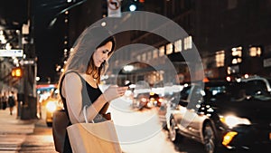 Young beautiful woman with shopping bags standing in traffic downtown in New York and using the smartphone.