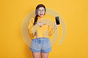 Young beautiful woman showing blank screen of smart phone with her index finger isolated over yellow background, keeping mouth