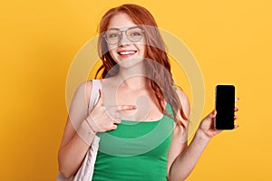 Young beautiful woman showing to camera blank screen of smart phone isolated over yellow background, being in good mood, red