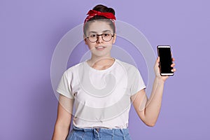 Young beautiful woman shows right in camera blank screen of smart phone isolated over lilac background, girl in white casual t