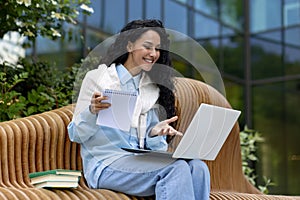 Young beautiful woman sits on a bench outside an office building, uses a laptop to watch a webinar course video, a