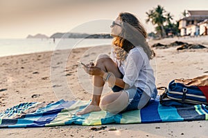Young beautiful woman sits on the sand of a beach with a smartphone in her hands during sunset