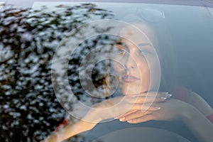 A young, beautiful woman sits at the wheel of the car and dreamingly watched through the glare of the front windshield.