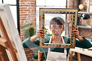 Young beautiful woman sitting at art studio with empty frame in shock face, looking skeptical and sarcastic, surprised with open
