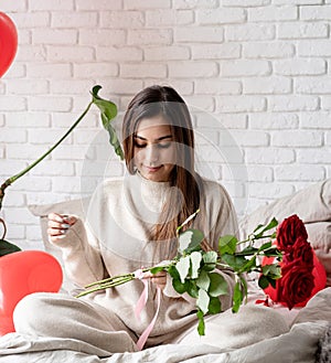 Young beautiful woman sitting in the bed celebrating valentine day holding red roses