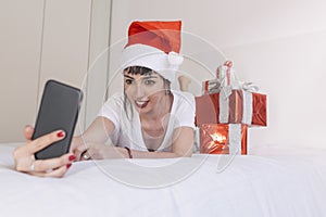 young beautiful woman sitting on bed and taking a selfie with mobile phone. Presents on bed. Wearing red santa hat. Christmas