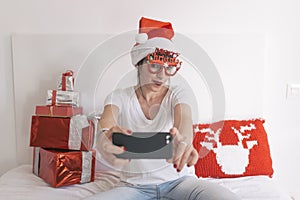 young beautiful woman sitting on bed and taking a selfie with mobile phone. Presents on bed. Wearing red santa hat and funny