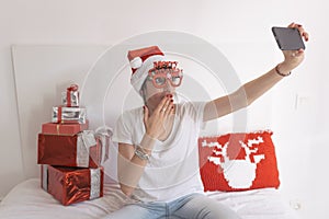 young beautiful woman sitting on bed and taking a selfie with mobile phone. Presents on bed. Wearing red santa hat and funny
