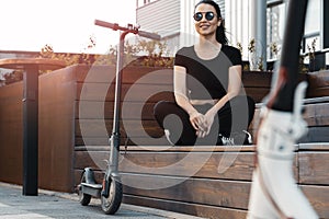 Young beautiful woman sitting on bench near electric kick scooter