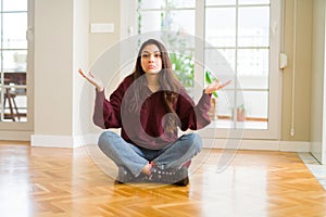 Young beautiful woman sitting on the floor at home clueless and confused expression with arms and hands raised