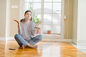 Young beautiful woman sitting on the floor at home clueless and confused expression with arms and hands raised