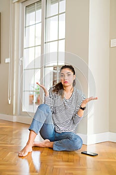 Young beautiful woman sitting on the floor at home clueless and confused expression with arms and hands raised