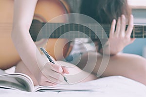 Young beautiful woman sitting on her bed in the bedroom holding guitar composing a song  and writing song in textbook ,musician,
