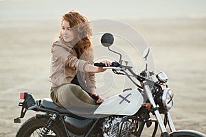 young beautiful woman sitting on her old cafe racer motorcycle in desert at sunset or sunrise