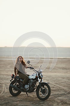 Young beautiful woman sitting on her old cafe racer motorcycle in desert at sunset or sunrise