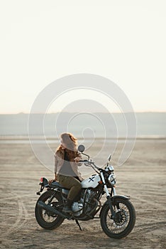 Young beautiful woman sitting on her old cafe racer motorcycle in desert at sunset or sunrise