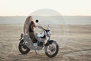 Young beautiful woman sitting on her old cafe racer motorcycle in desert at sunset or sunrise