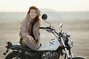 Young beautiful woman sitting on her old cafe racer motorcycle in desert at sunset or sunrise